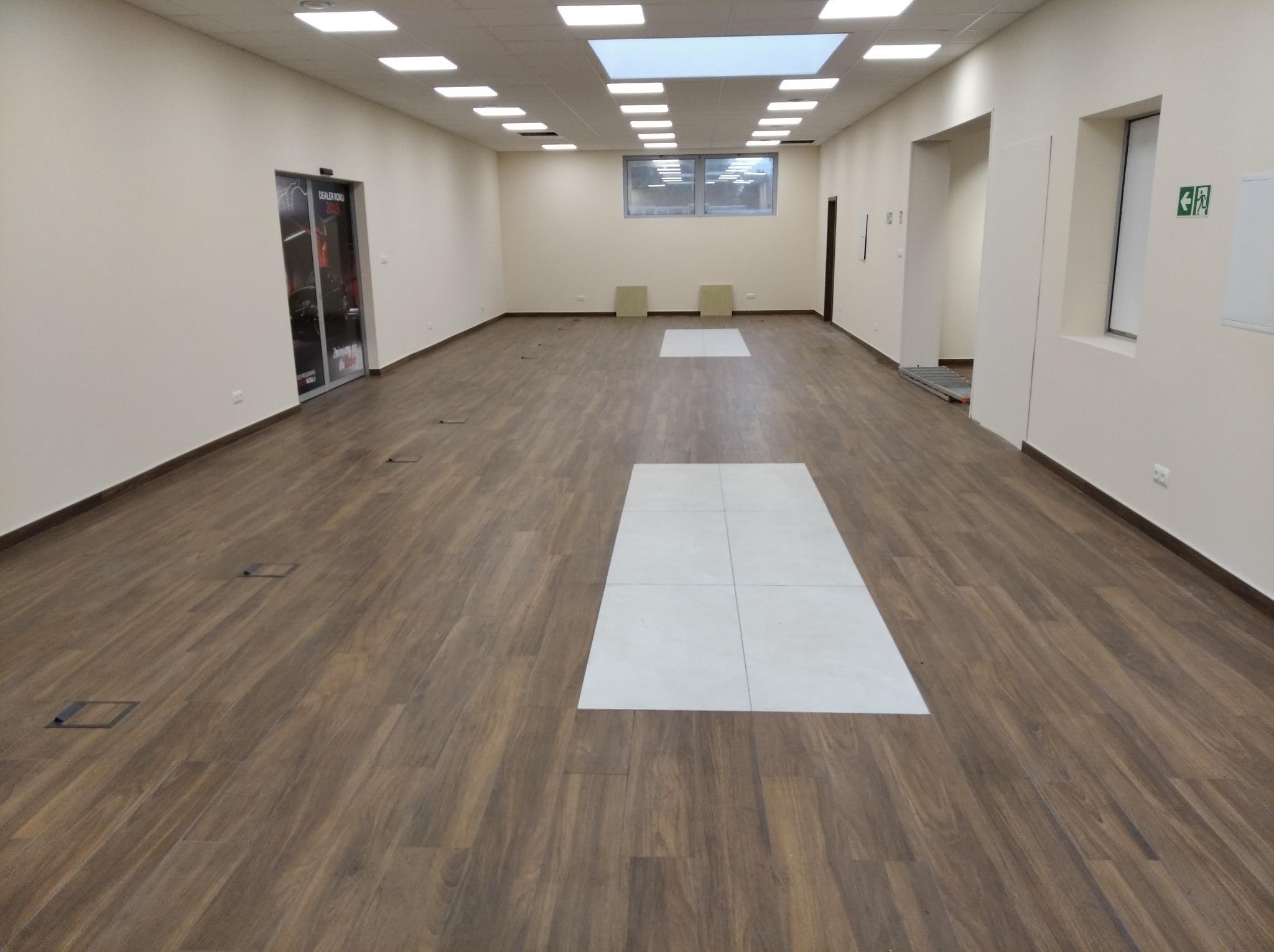 Długi, pusty korytarz wykończony panelami podłogowymi imitującymi drewno, z wstawkami z jasnych płytek, oświetlony szeregiem lamp sufitowych i naturalnym światłem z okna na końcu korytarza.