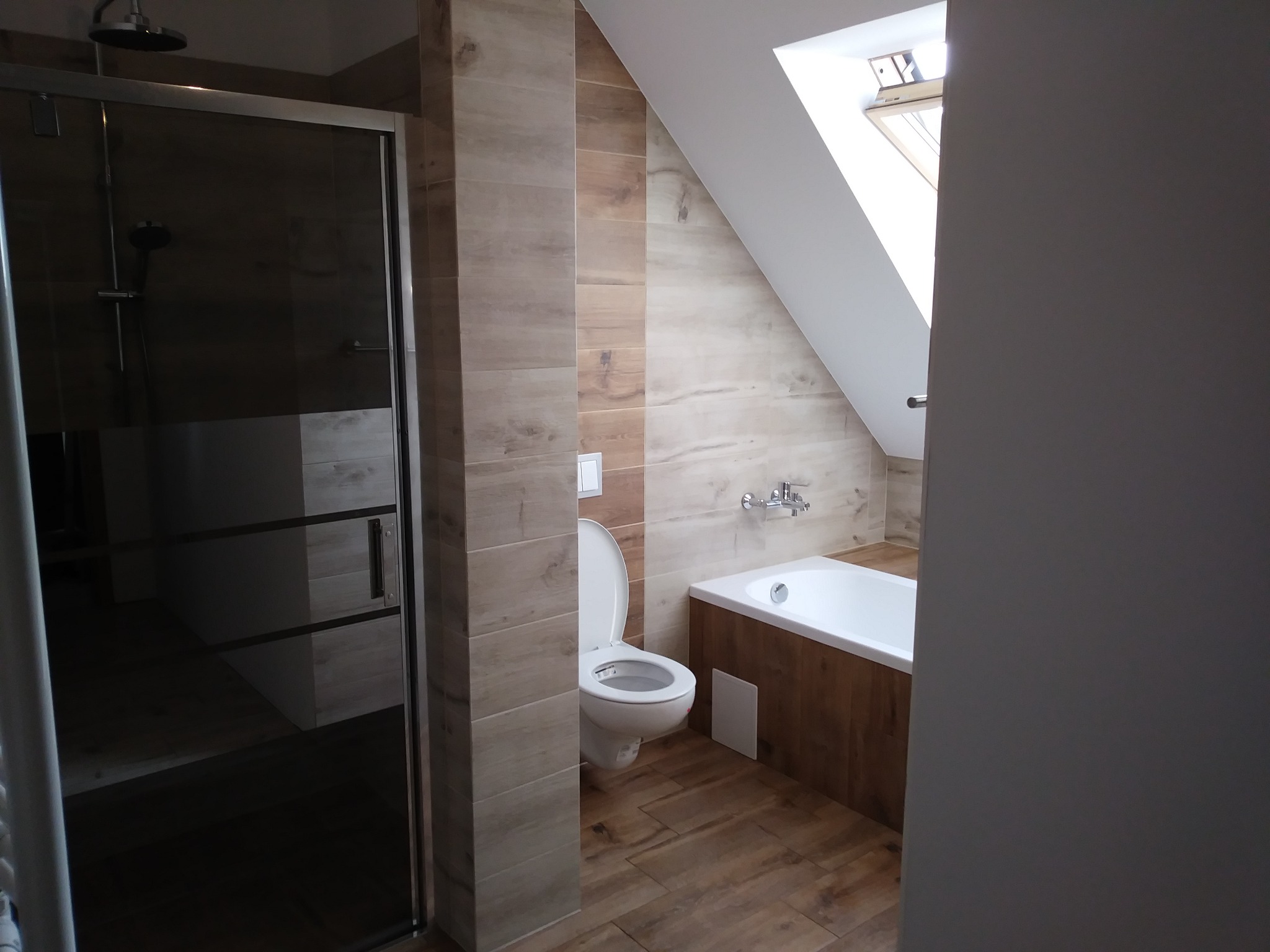 Jasna łazienka na poddaszu z drewnopodobnymi płytkami, kabiną prysznicową ze szklanymi drzwiami i białą wanną z obudową z drewnopodobnych paneli.