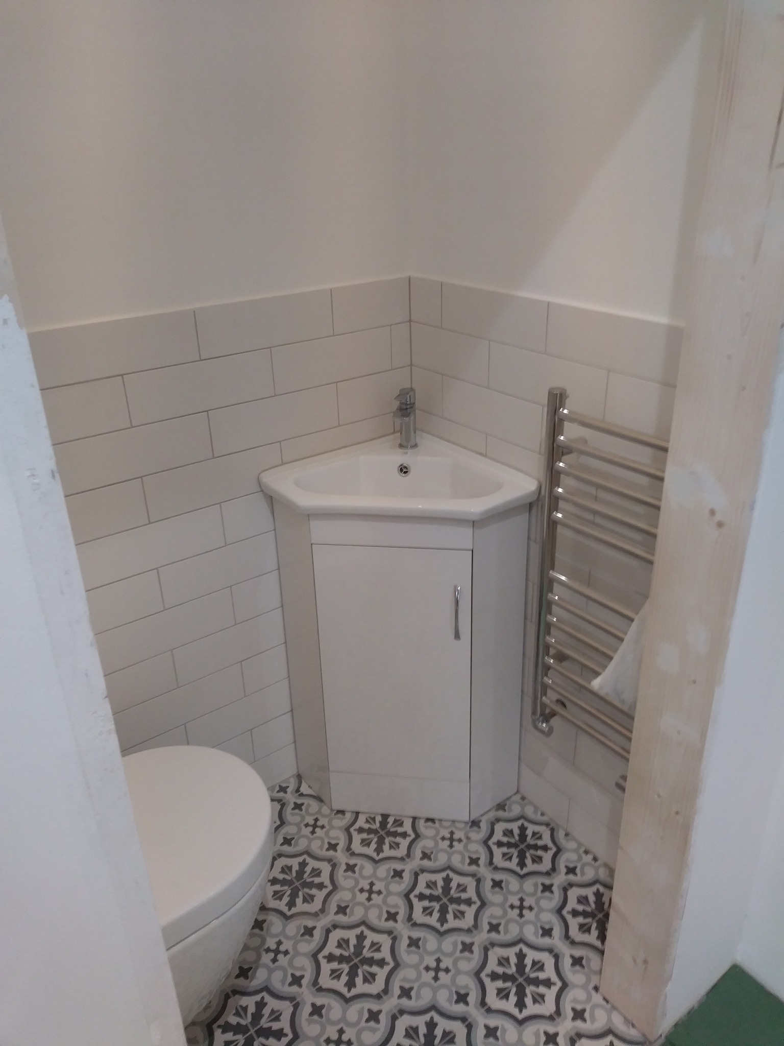 Narożna umywalka z szafką w małej łazience, białe płytki na ścianach, wzorzyste szaro-czarne płytki podłogowe, grzejnik drabinkowy po prawej stronie.