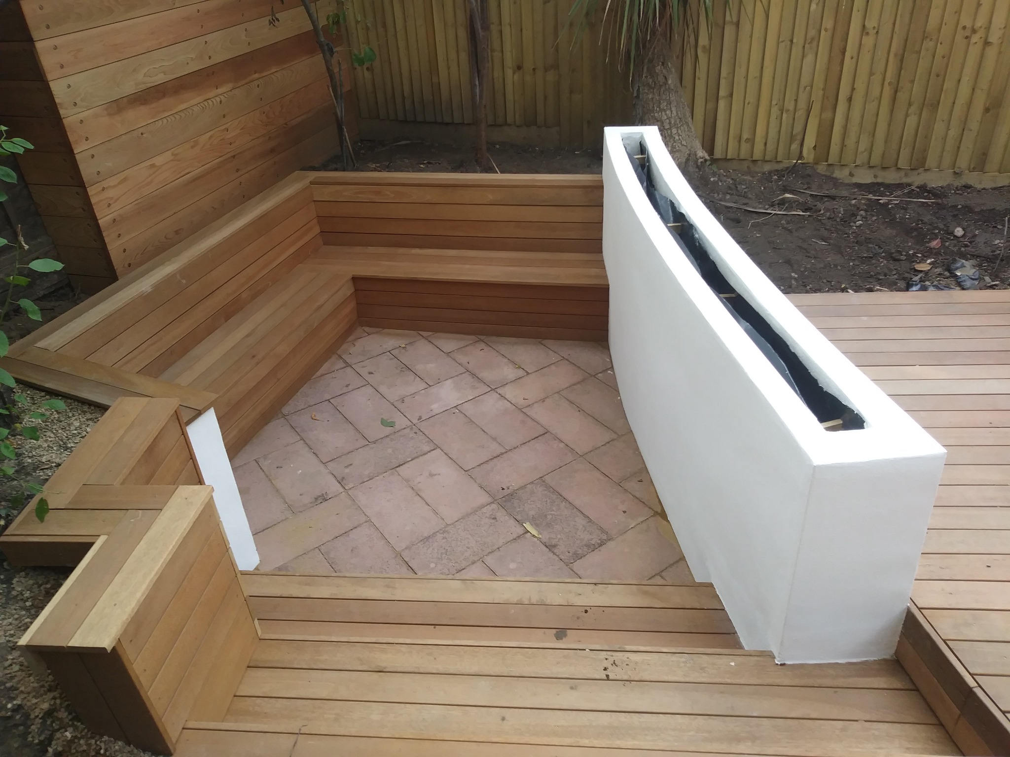 Drewniany taras z wbudowanymi siedziskami i białą donicą, wyłożony ceglaną kostką, otoczony drewnianym płotem i drzewami.