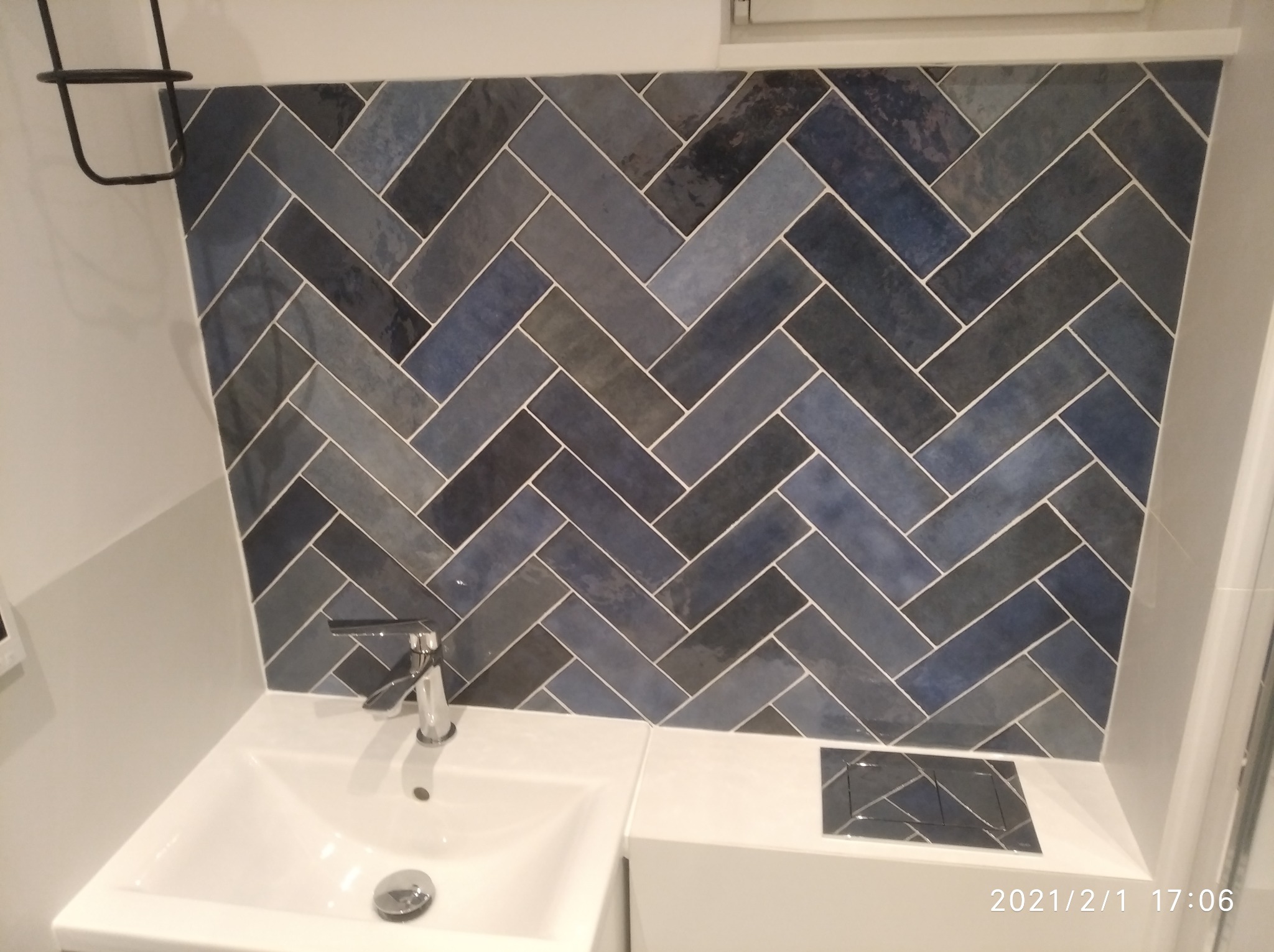 Błękitno-szara ściana z płytek ułożonych w jodełkę nad białą umywalką z chromowaną baterią w łazience.