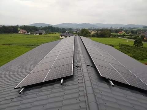 Panele słoneczne zamontowane na ciemnoszarym dachu z dachówki ceramicznej, widok z góry na tle zielonych pól i wzgórz w pochmurny dzień.