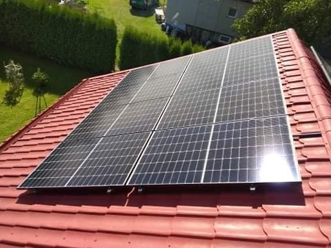 Panele fotowoltaiczne zamontowane na czerwonym, spadzistym dachu, z widocznym ogrodem i budynkiem w tle.