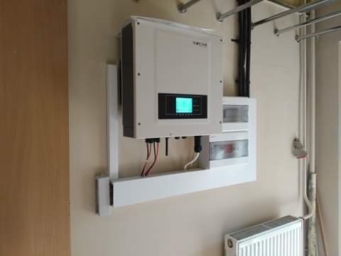 Biały falownik z wyświetlaczem i okablowaniem zamontowany na ścianie, widoczna skrzynka bezpiecznikowa i rury instalacyjne.