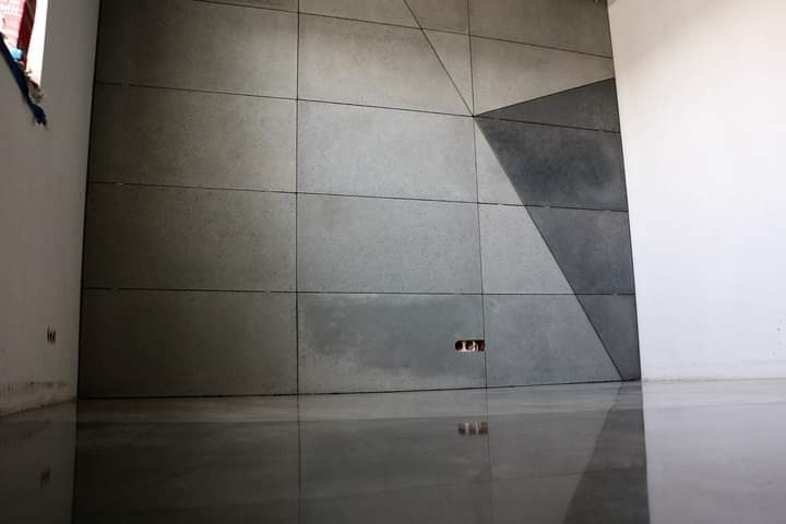 Ściana wyłożona szarymi, betonowymi płytami z geometrycznym wzorem, odbijająca się w lśniącej, polerowanej podłodze. Odsłonięte gniazdko elektryczne w dolnej części ściany. Fragment białej ściany...