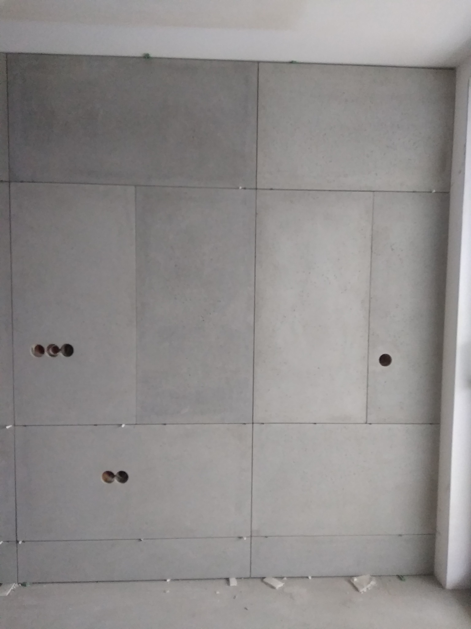 Ściana wykończona płytami o szarym, betonowym wyglądzie, z widocznymi otworami na gniazdka elektryczne i zielonymi elementami mocującymi w szczelinach między płytami.