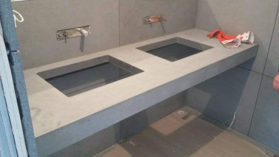 Surowy betonowy blat z dwoma wyciętymi otworami na umywalki, widoczne instalacje wodne w ścianie, czerwona ściereczka leży na blacie, szare płytki na ścianach i podłodze, w trakcie montażu.