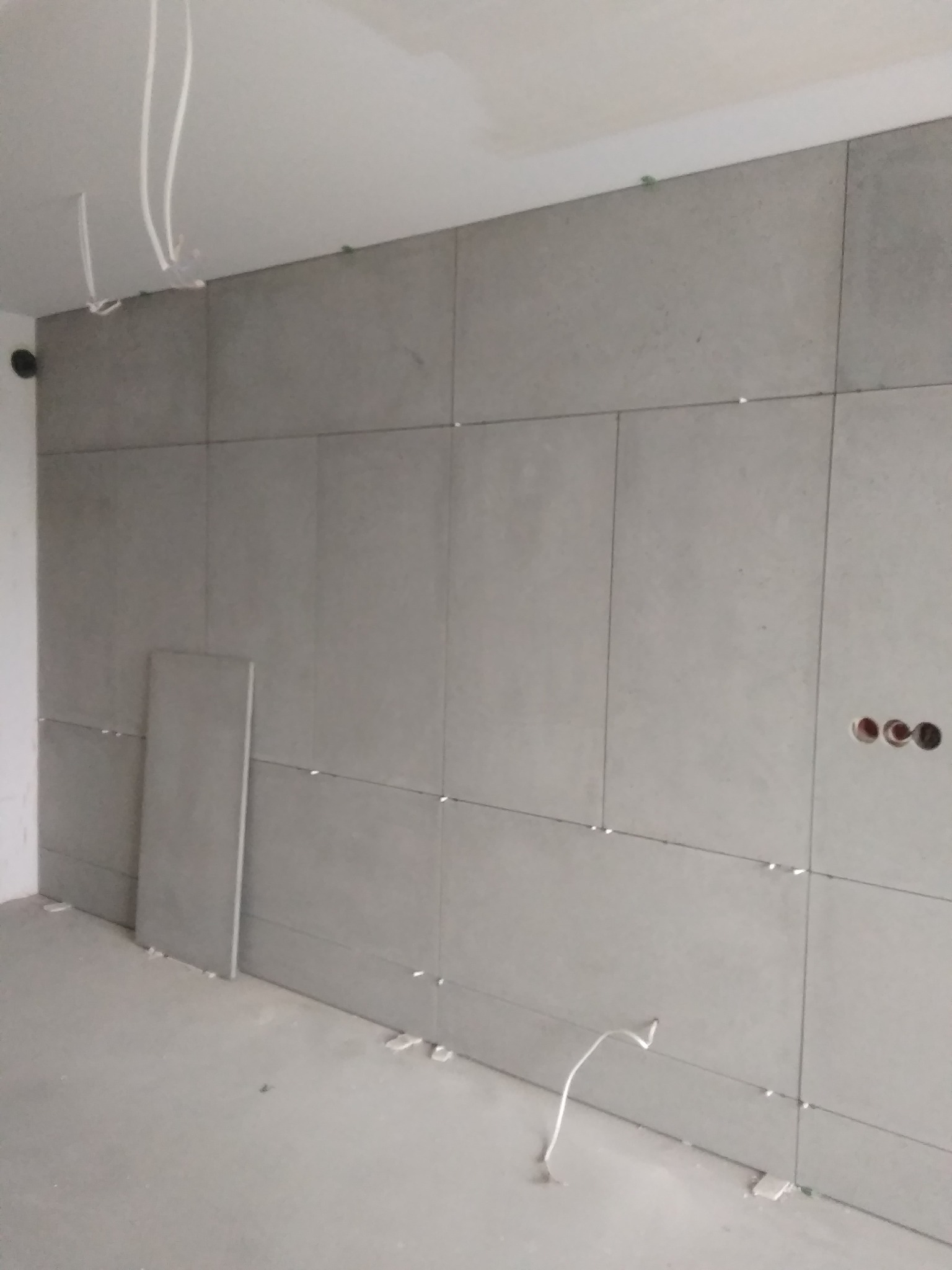 Ściana w trakcie układania płytek w odcieniach szarości, widoczne krzyżyki dystansowe, przewody elektryczne wystające z sufitu i puszki elektryczne w ścianie.