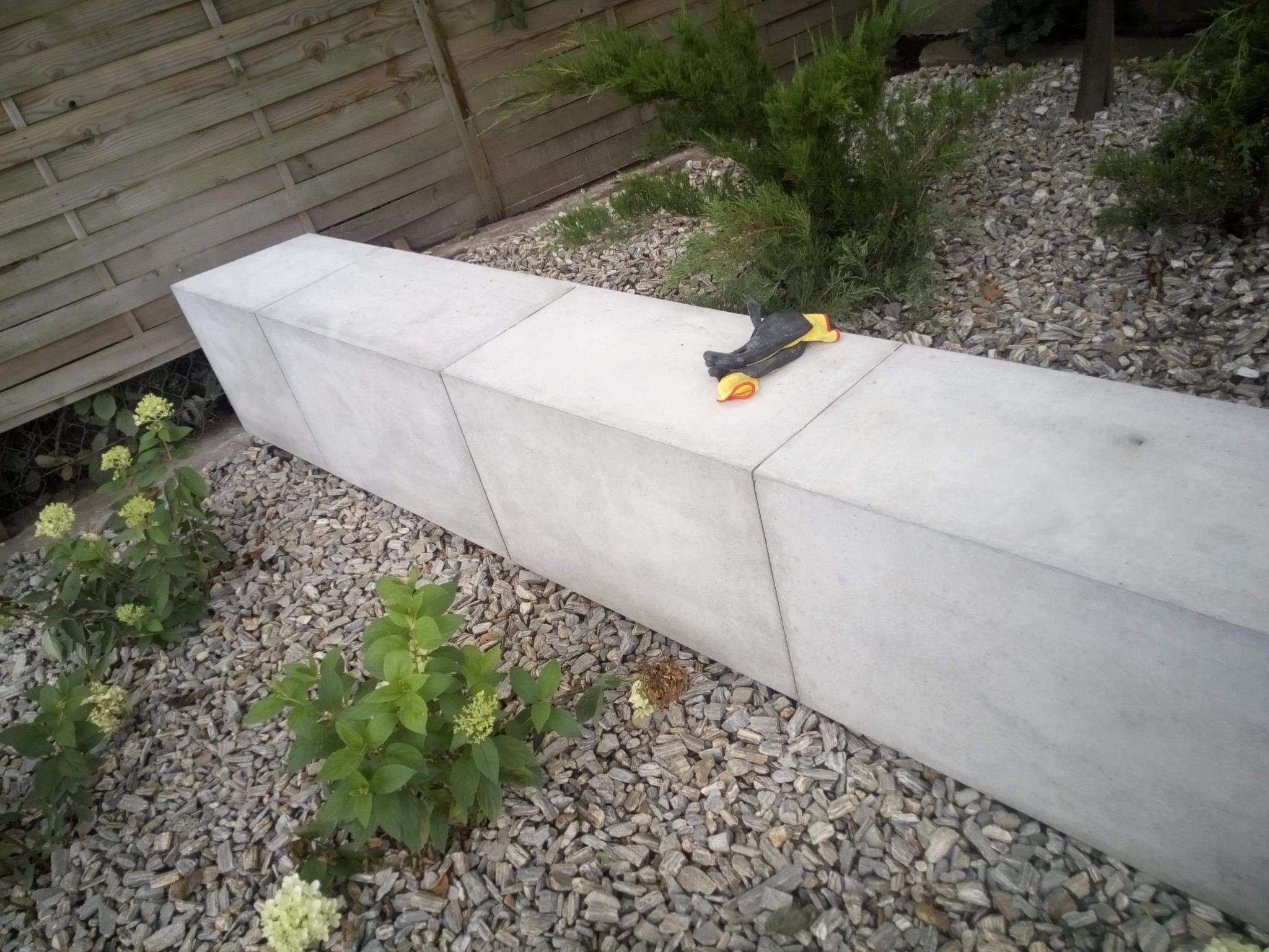 Betonowy murek ogrodowy zbudowany z kilku segmentów, otoczony żwirem i roślinami, z roboczymi rękawicami leżącymi na górze.