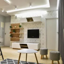 Jasne wnętrze pokoju dziennego z geometryczną tapetą, drewnianymi półkami z dekoracjami, białymi meblami i szarymi zasłonami.