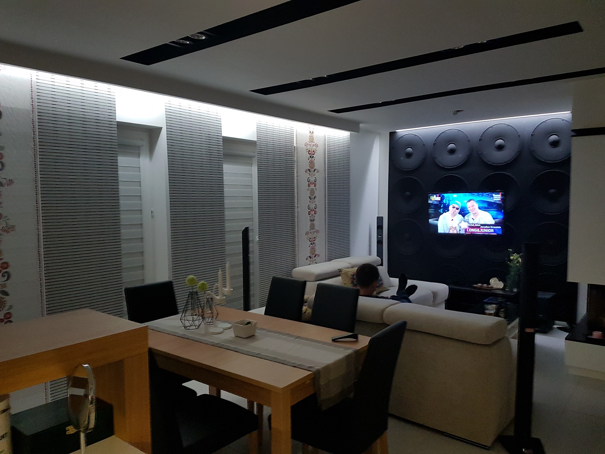 Nowoczesne wnętrze salonu z drewnianym stołem jadalnym, szarymi zasłonami panelowymi z motywem kwiatowym, czarną ścianą dekoracyjną z okrągłymi elementami i telewizorem, oraz beżową sofą.