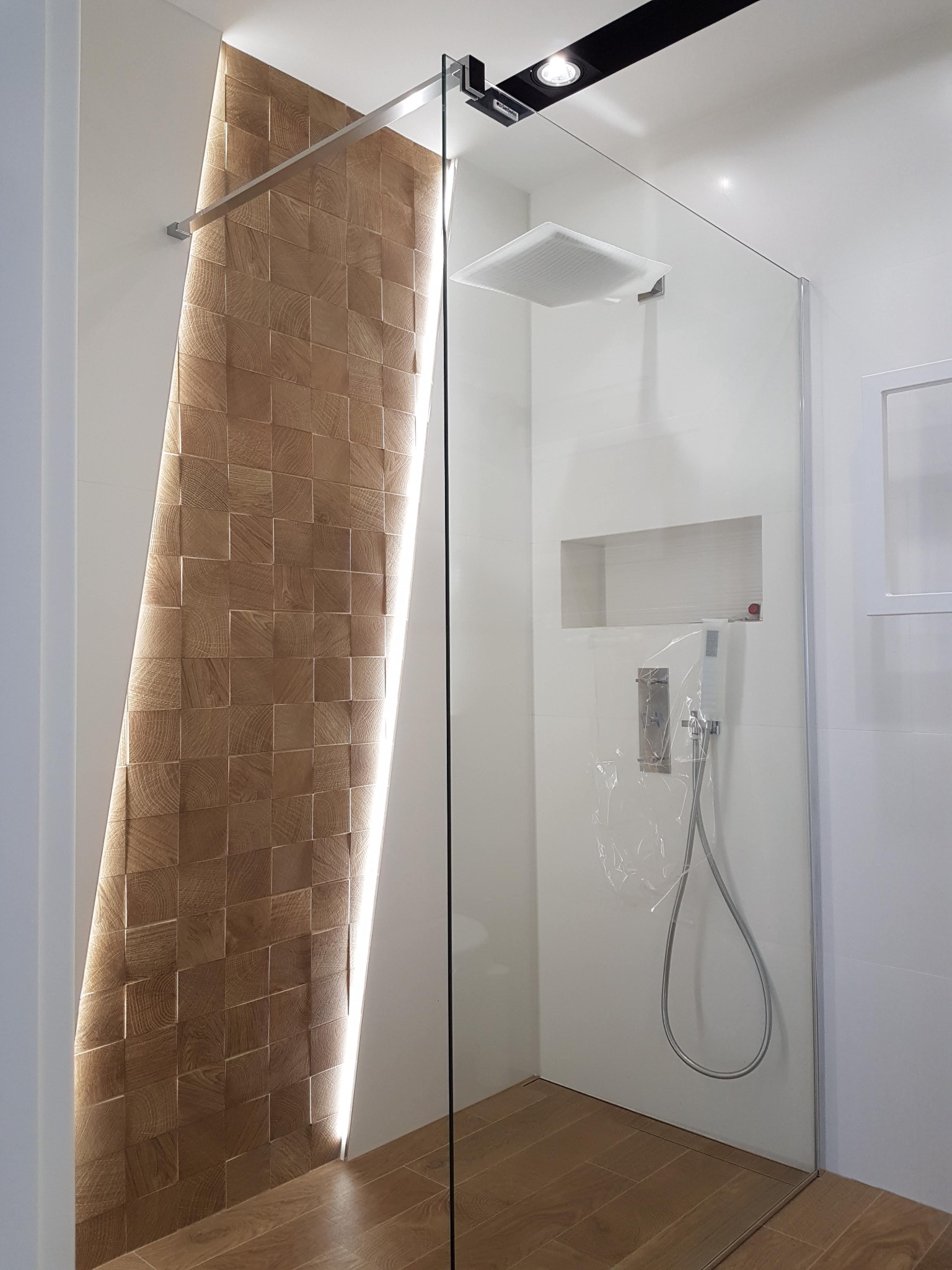 Nowoczesna kabina prysznicowa z drewnianą mozaiką ścienną podświetloną od tyłu, szklana ściana kabiny, armatura zabezpieczona folią ochronną, płytki podłogowe imitujące drewno.