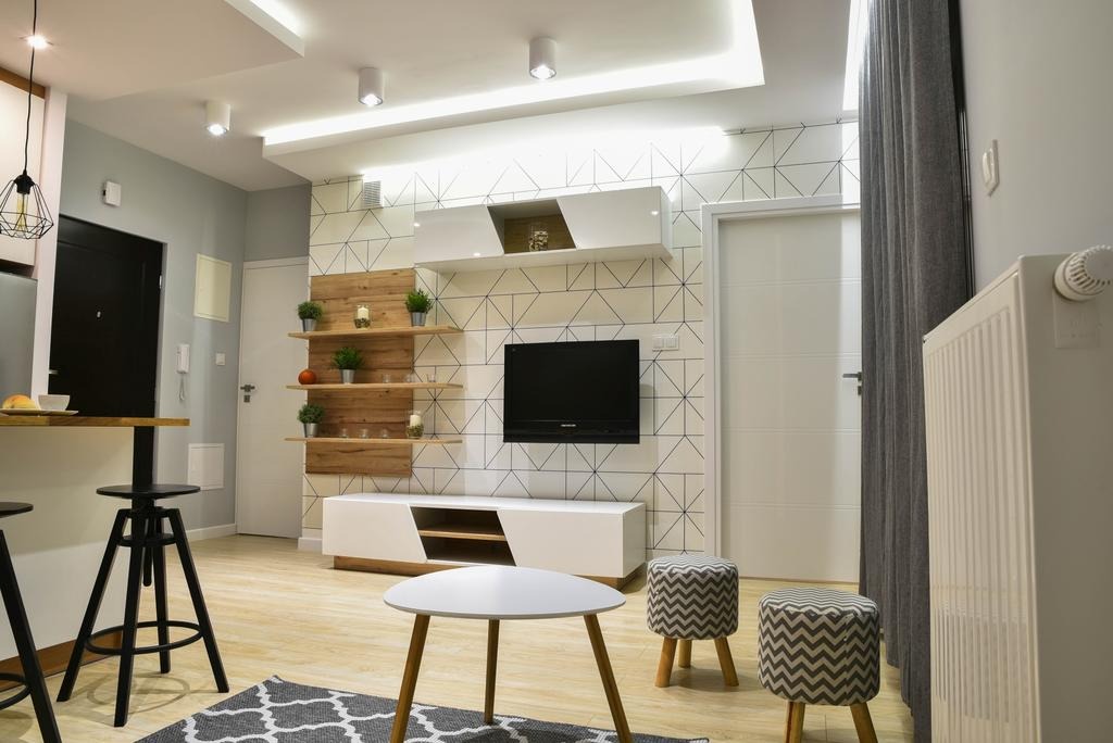 Jasne wnętrze pokoju dziennego z geometryczną tapetą, drewnianymi półkami z dekoracjami, białymi meblami i szarymi zasłonami.