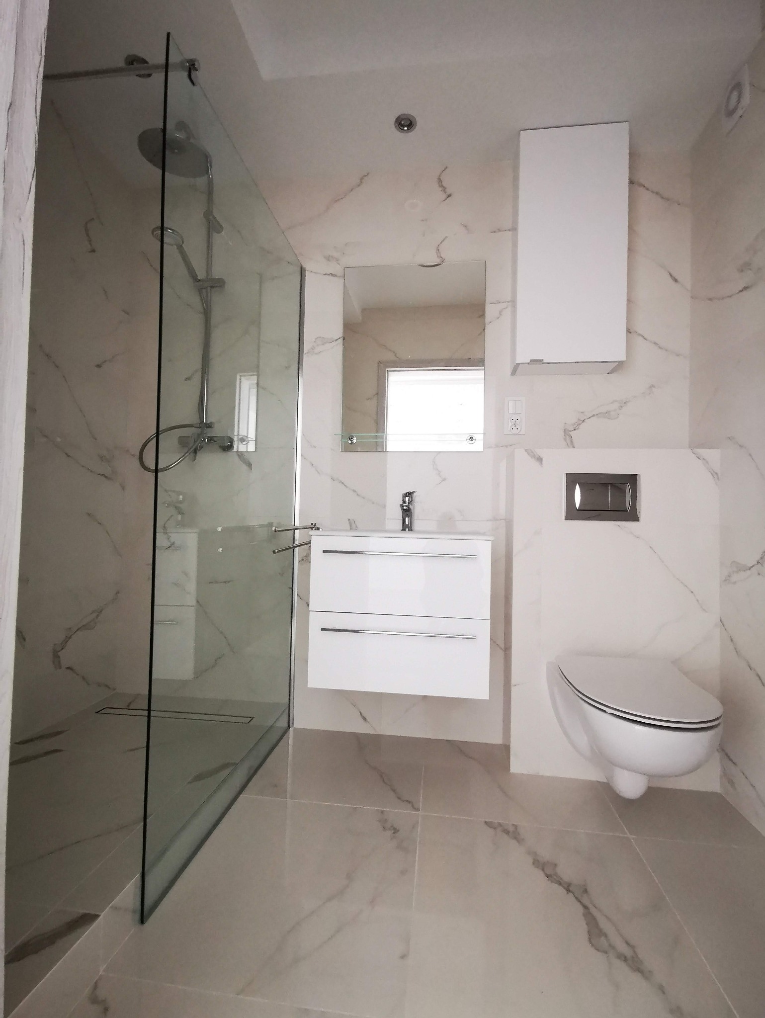 Nowoczesna łazienka z kabiną prysznicową typu walk-in, białą umywalką z szafką, wiszącą toaletą i marmurowymi płytkami na ścianach i podłodze.