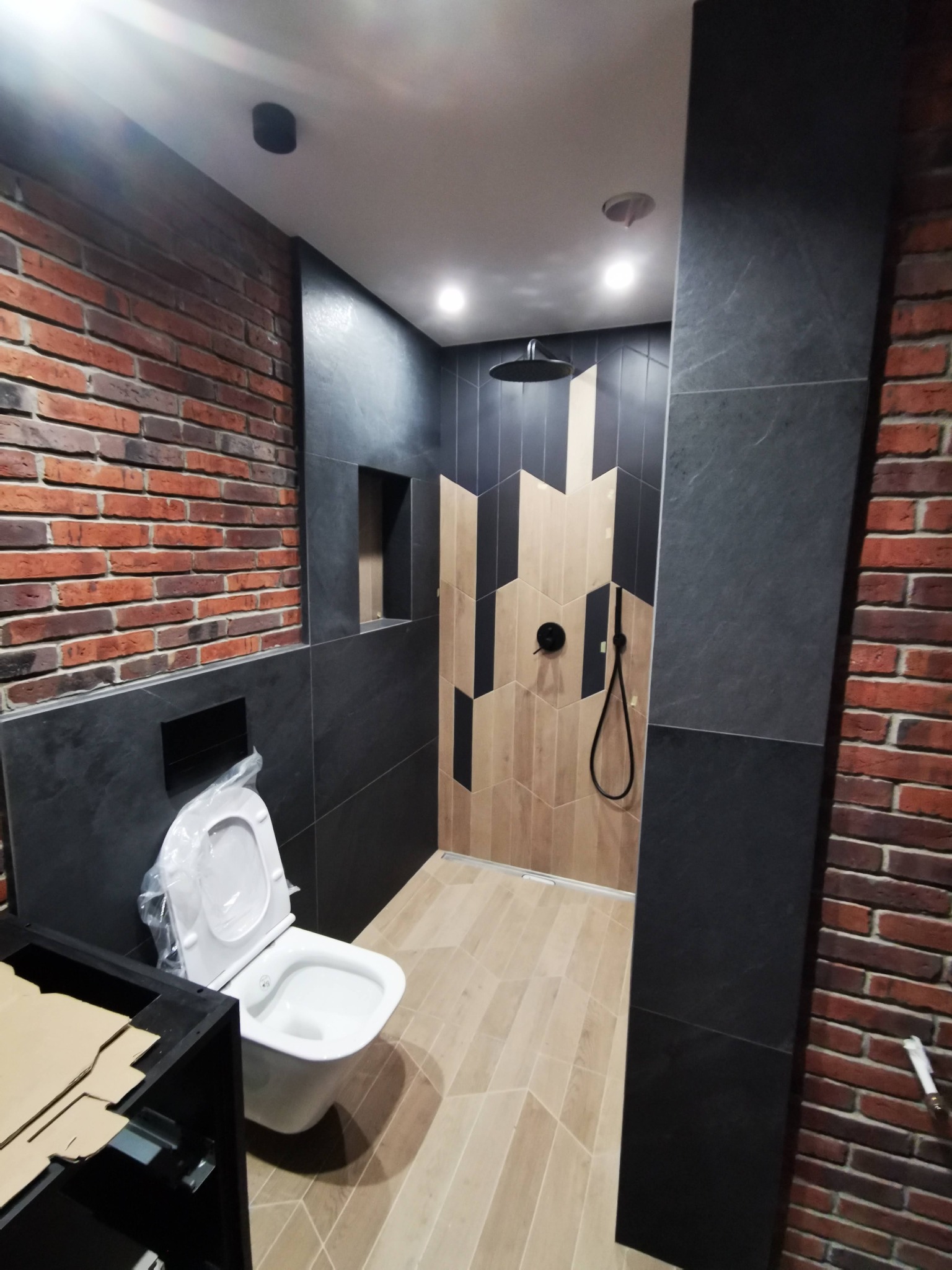 Nowoczesna łazienka w Rzeszowie w trakcie remontu: ściana z cegły, czarne płytki, prysznic z deszczownicą i toaleta z częściowo zamontowaną szafką.