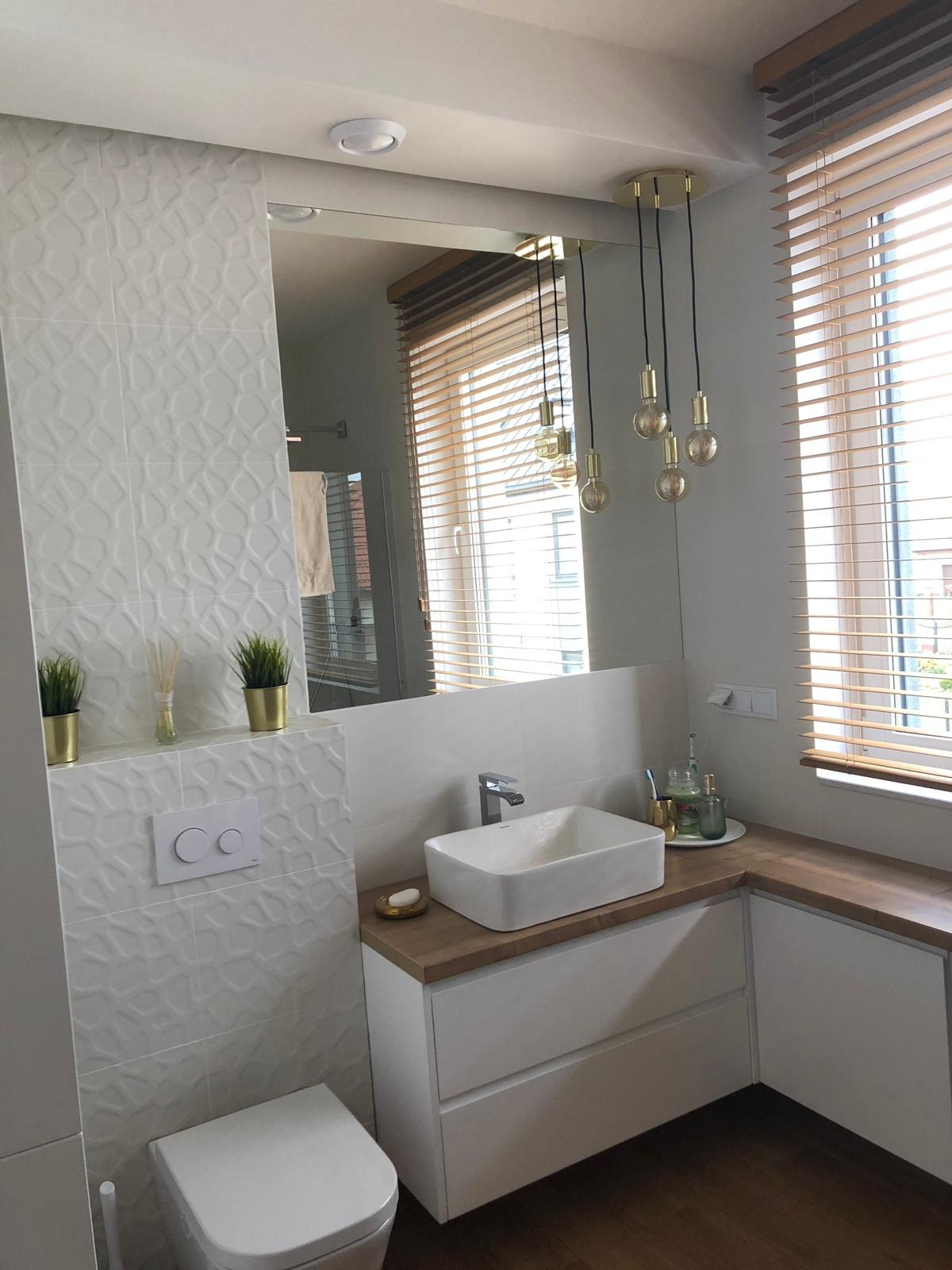 Nowoczesna łazienka z białą umywalką nablatową, armaturą, białą toaletą, płytkami 3D na ścianie, drewnianym blatem i żarówkami dekoracyjnymi wiszącymi nad lustrem.
