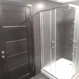 Nowoczesna łazienka z kabiną prysznicową z matowego szkła i ciemnymi płytkami oraz ciemnymi drzwiami z poziomymi, jasnymi wstawkami.