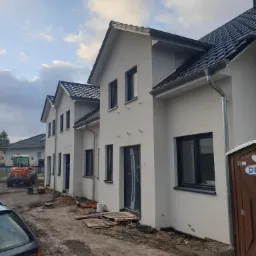 Nowo wybudowane, otynkowane domy szeregowe we Wrocławiu, z czarną dachówką i ramami okiennymi, widoczne ślady prac ziemnych i lekki sprzęt budowlany.