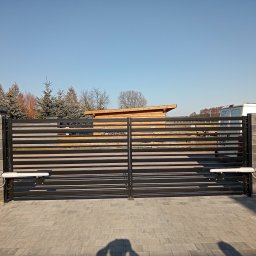 Instancje elektryczne - Nowoczesna czarna brama dwuskrzydłowa z poziomymi przęsłami, z białymi siłownikami automatyki, na słupkach z szarych bloczków, przy brukowanym podjeździe.