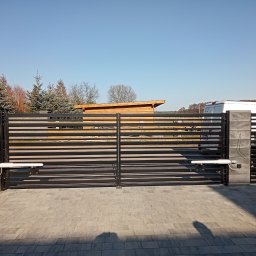 Instancje elektryczne - Nowoczesna czarna brama dwuskrzydłowa z poziomymi lamelami i dwoma białymi siłownikami automatycznymi, osadzona w szarych słupkach z kostki. Przed nią szara kostka brukowa, w tle drzewa i biały van.