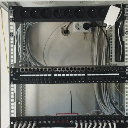 Instancje elektryczne - Szafa rackowa z listwami zasilającymi, dwoma patch panelami Cat.6 i Cat.5e, modułem antenowym. Uporządkowane okablowanie sieciowe i elektryczne na tle surowej ściany.