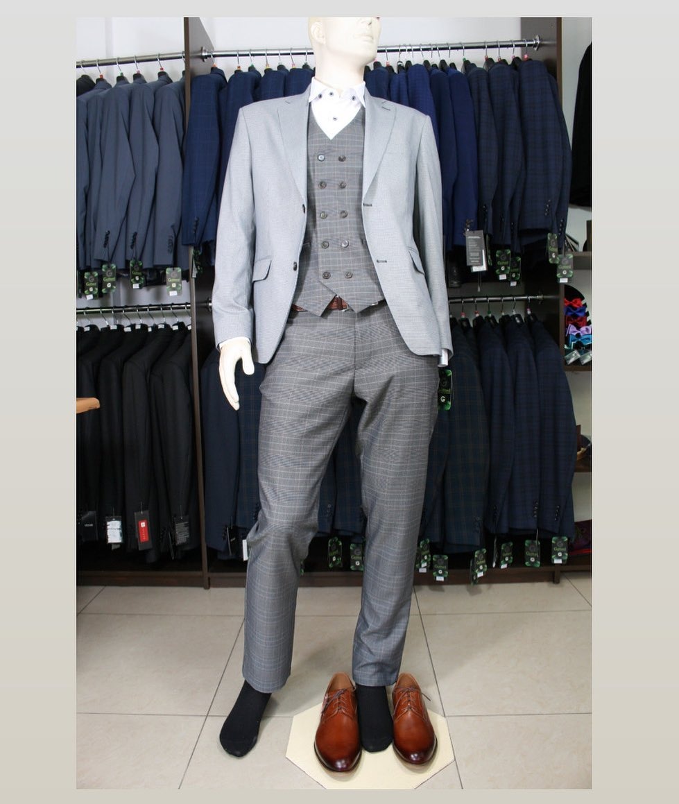 Manekin w szarym garniturze w kratę z kamizelką, brązowe buty na podkładce, w tle wieszaki z garniturami.