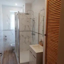 Nowoczesna łazienka z kabiną prysznicową typu walk-in, umywalką na szafce, toaletą i drewnianymi drzwiami z lamelami, wykończona płytkami imitującymi marmur.