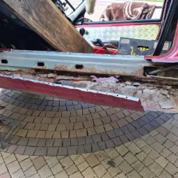 Czerwony samochód na podnośniku z odsłoniętym, skorodowanym progiem, częściowo zabezpieczonym nowym elementem blacharskim z zaznaczonymi liniami cięcia.