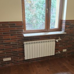 Pan Remoncik - Ściana wyłożona ceglastą płytką klinkierową do połowy wysokości, z oknem i grzejnikiem. Widoczne gniazdka elektryczne i drewniana podłoga.