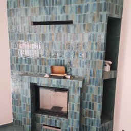 Pan Remoncik - Turkusowy kominek obłożony błyszczącymi, pionowo ułożonymi płytkami. Widoczne akcesoria na półce kominka i wnęka obok. Podłoga w kolorze płytek.