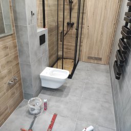 Pan Remoncik - Nowoczesna łazienka z wiszącą toaletą, kabiną prysznicową z czarnymi ramami i grzejnikiem drabinkowym. Szare płytki i drewnopodobne akcenty. Widoczne narzędzia i materiały.