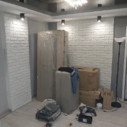Wnętrze w trakcie remontu: biała ściana z cegły, zapakowane meble, kartony, lampa podłogowa i narzędzia glazurnicze na podłodze.