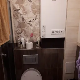 Biały piec gazowy Beretta na ścianie łazienki z beżowymi i brązowymi płytkami, nad toaletą i półką z kosmetykami.