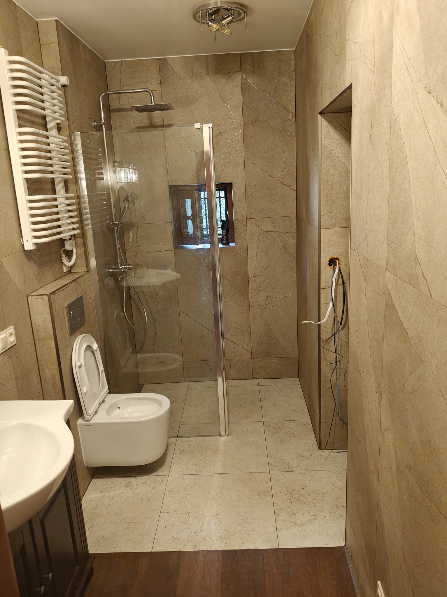 Wykończona łazienka z kabiną prysznicową typu walk-in, wiszącą toaletą i grzejnikiem drabinkowym. Płytki imitujące marmur na ścianach i podłodze. Widoczne kable elektryczne.