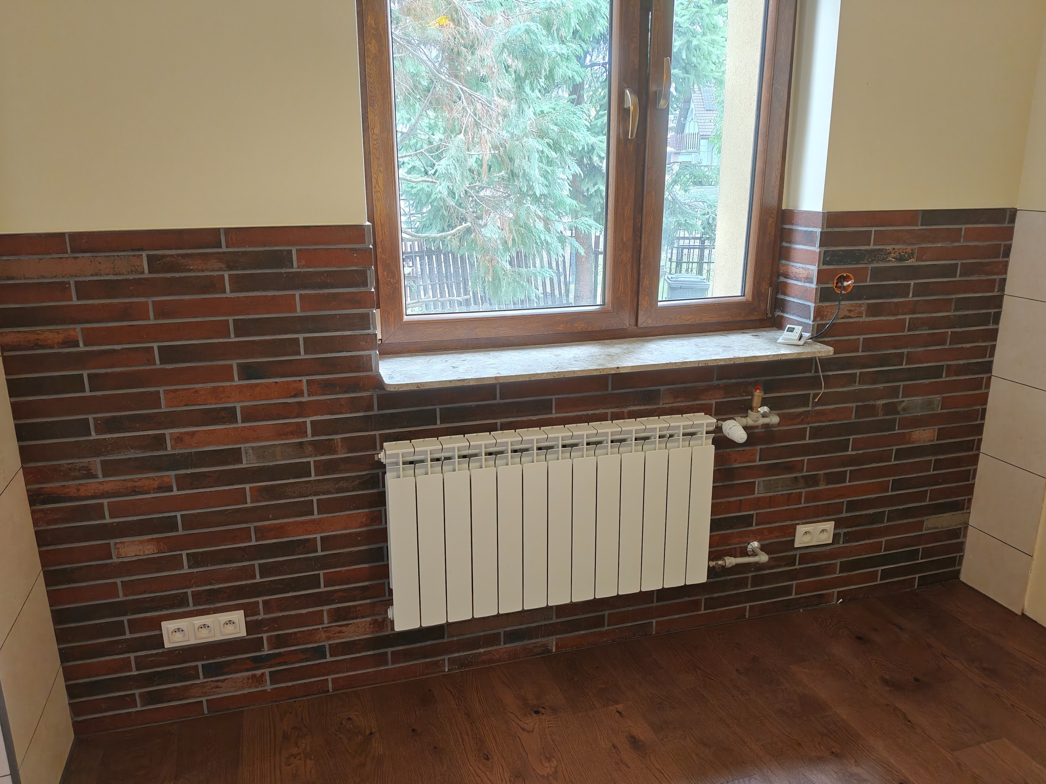 Ściana wyłożona ceglastą płytką klinkierową do połowy wysokości, z oknem i grzejnikiem. Widoczne gniazdka elektryczne i drewniana podłoga.