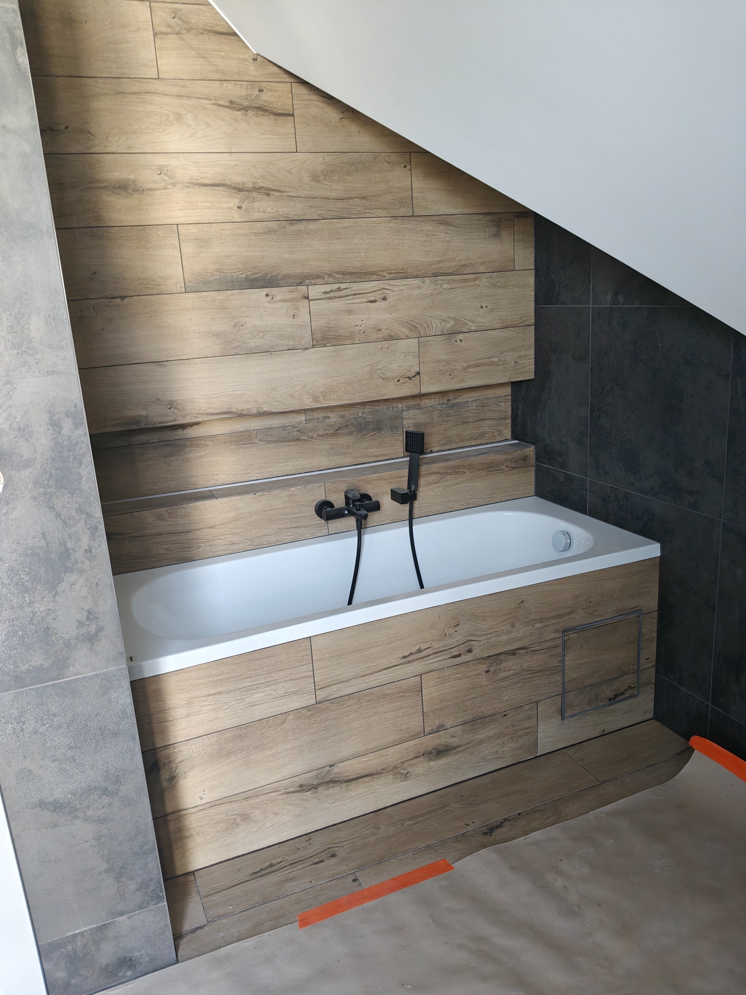 Nowoczesna łazienka na poddaszu z wanną obudowaną płytkami imitującymi drewno i czarnymi płytkami na ścianie. Widoczna armatura w kolorze czarnym.