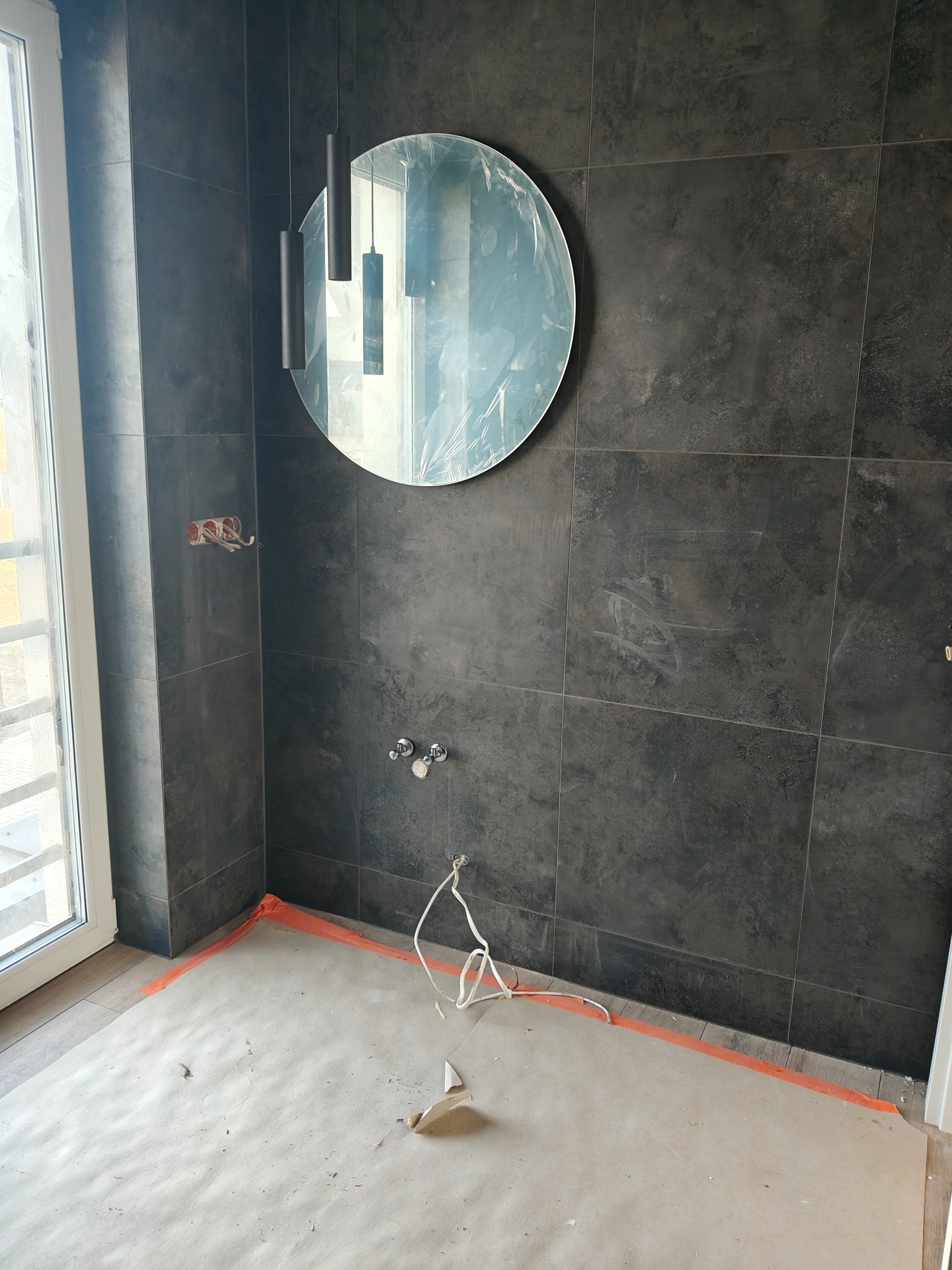 Łazienka w trakcie wykończenia: ciemne płytki na ścianach, okrągłe lustro z folią ochronną, wiszące lampy, przygotowane instalacje, zabezpieczona podłoga.