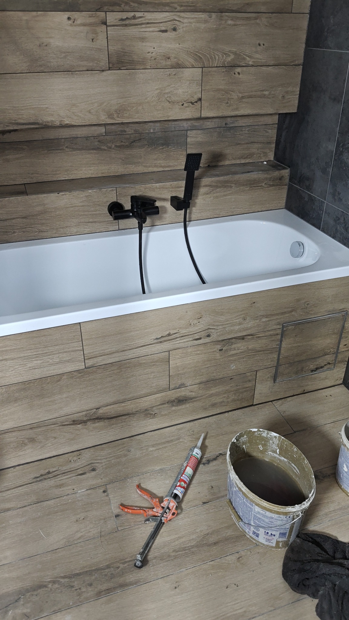 Wykończona łazienka z wanną obudowaną płytkami imitującymi drewno, czarna armatura, pistolet do silikonu i wiadro na podłodze. Detal z bliska.
