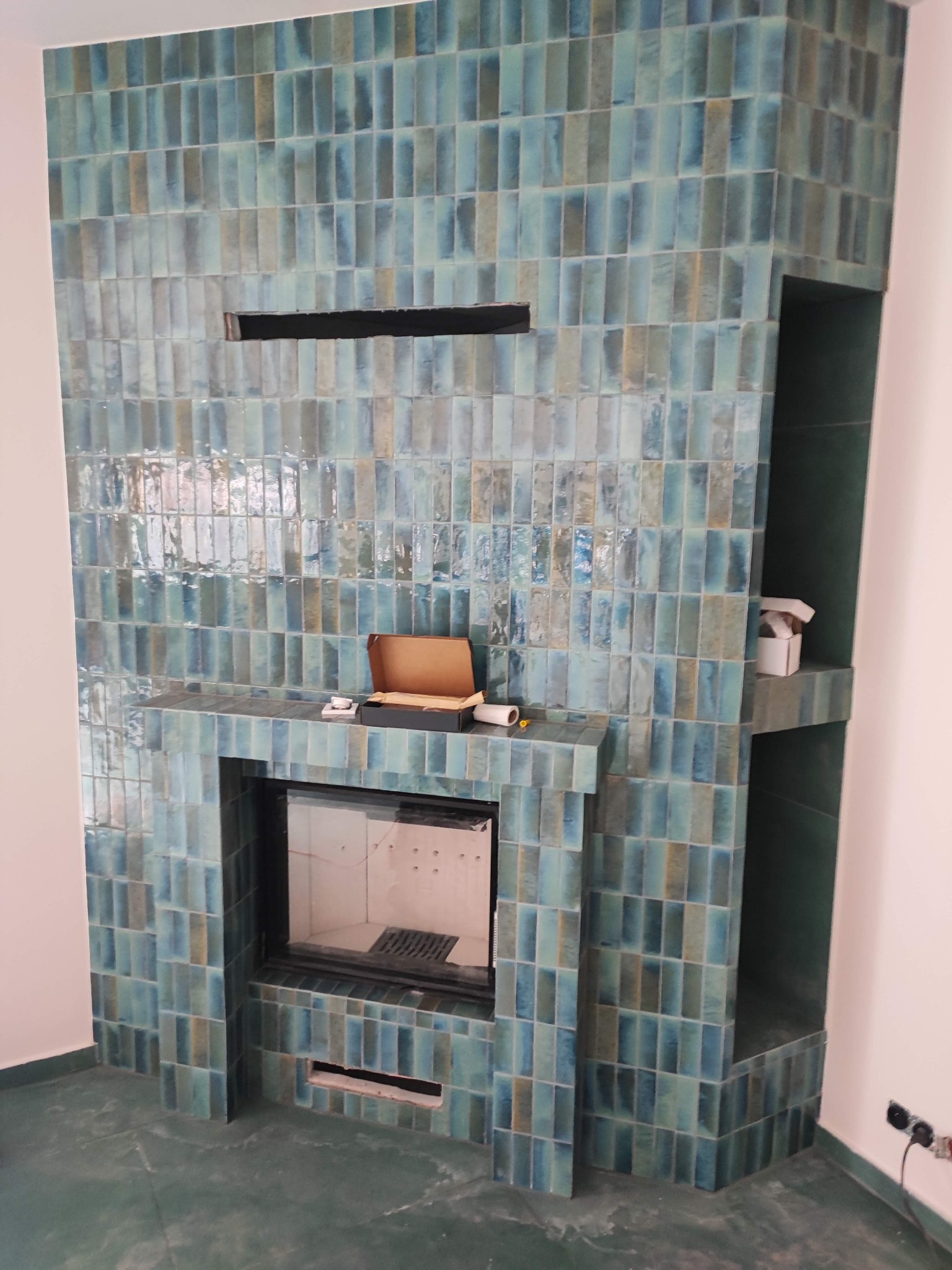 Turkusowy kominek obłożony błyszczącymi, pionowo ułożonymi płytkami. Widoczne akcesoria na półce kominka i wnęka obok. Podłoga w kolorze płytek.