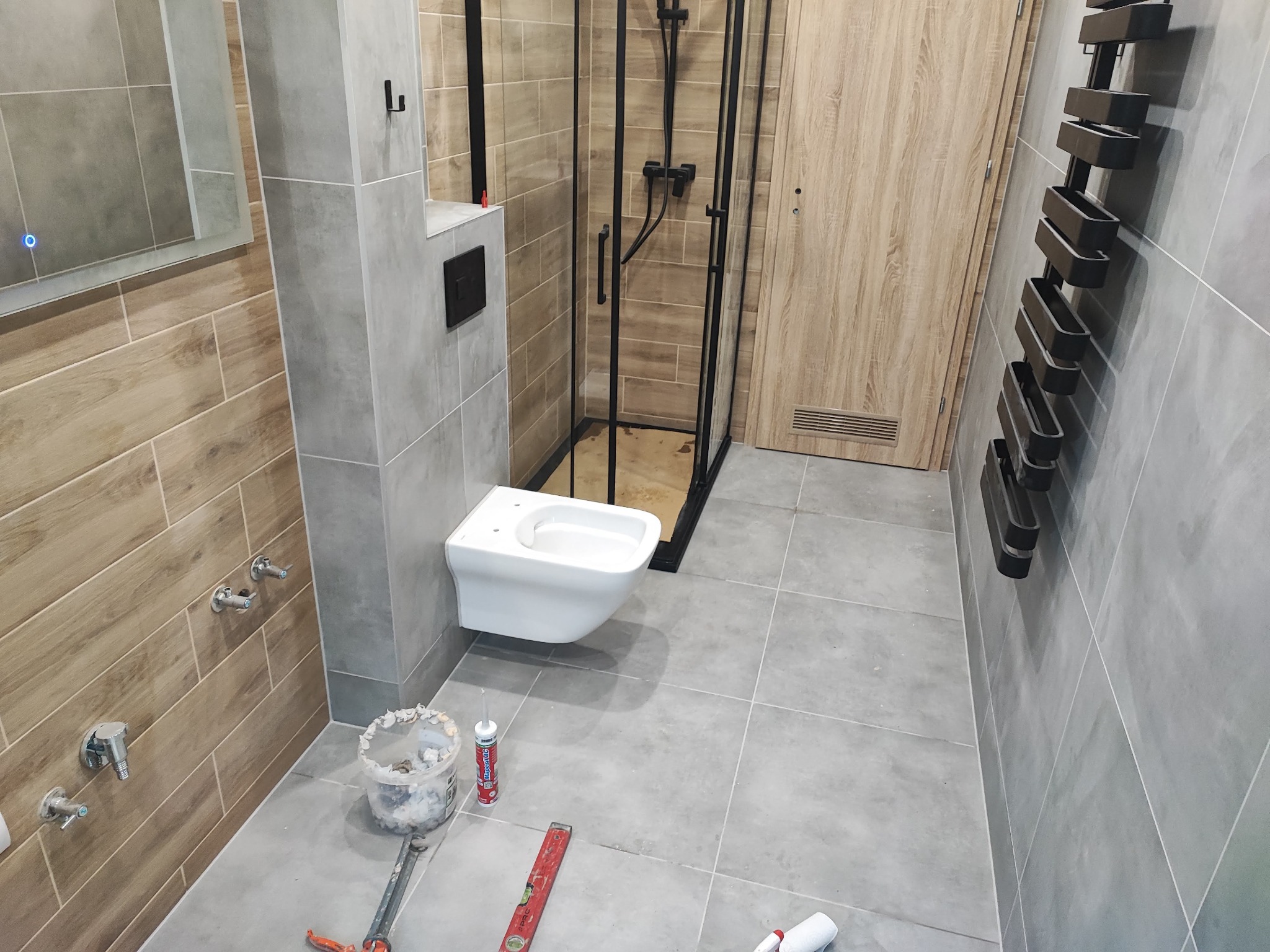 Nowoczesna łazienka z wiszącą toaletą, kabiną prysznicową z czarnymi ramami i grzejnikiem drabinkowym. Szare płytki i drewnopodobne akcenty. Widoczne narzędzia i materiały.