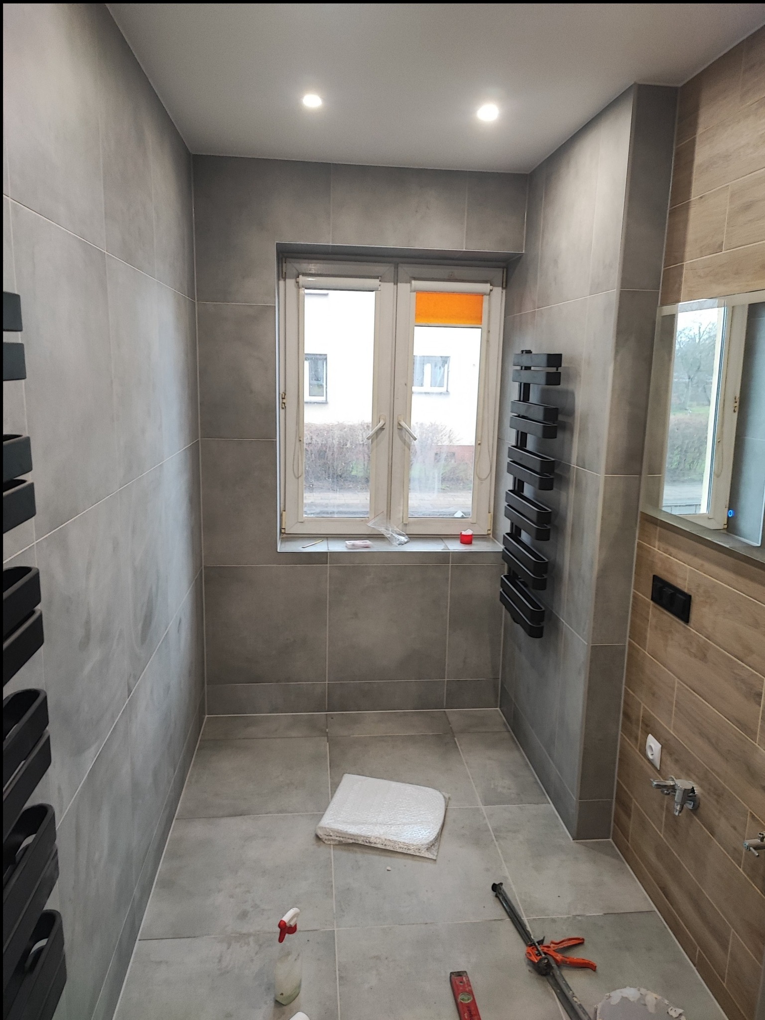 Wykończona łazienka w szarościach z elementami drewna. Widoczne płytki na ścianach i podłodze, grzejnik drabinkowy, okno z pomarańczową roletą i narzędzia glazurnicze.