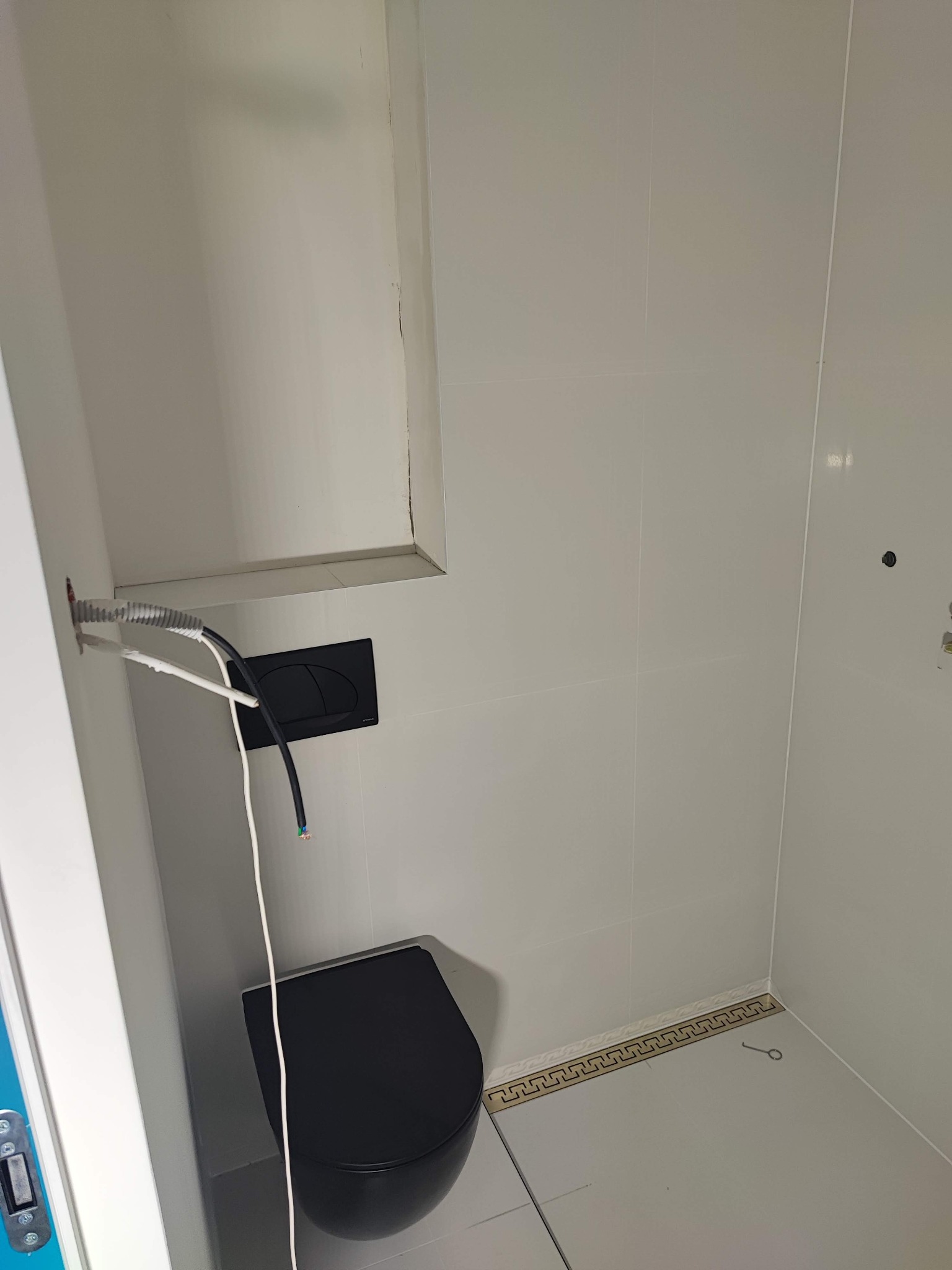 Biała łazienka z czarną toaletą podtynkową i widocznymi przewodami elektrycznymi. Dekoracyjny odpływ liniowy przy podłodze.