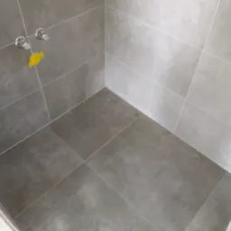 Fragment łazienki z wyłożonymi szarymi płytkami podłogowymi i ściennymi, widoczne przyłącza wodne zabezpieczone żółtą folią ochronną.