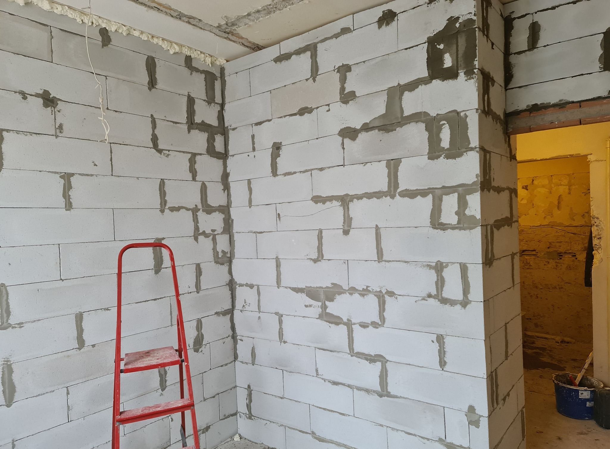Wnętrze w trakcie budowy z białych bloczków betonowych, widoczna czerwona drabina i wiadro z zaprawą, pianka montażowa w szczelinach przy suficie.