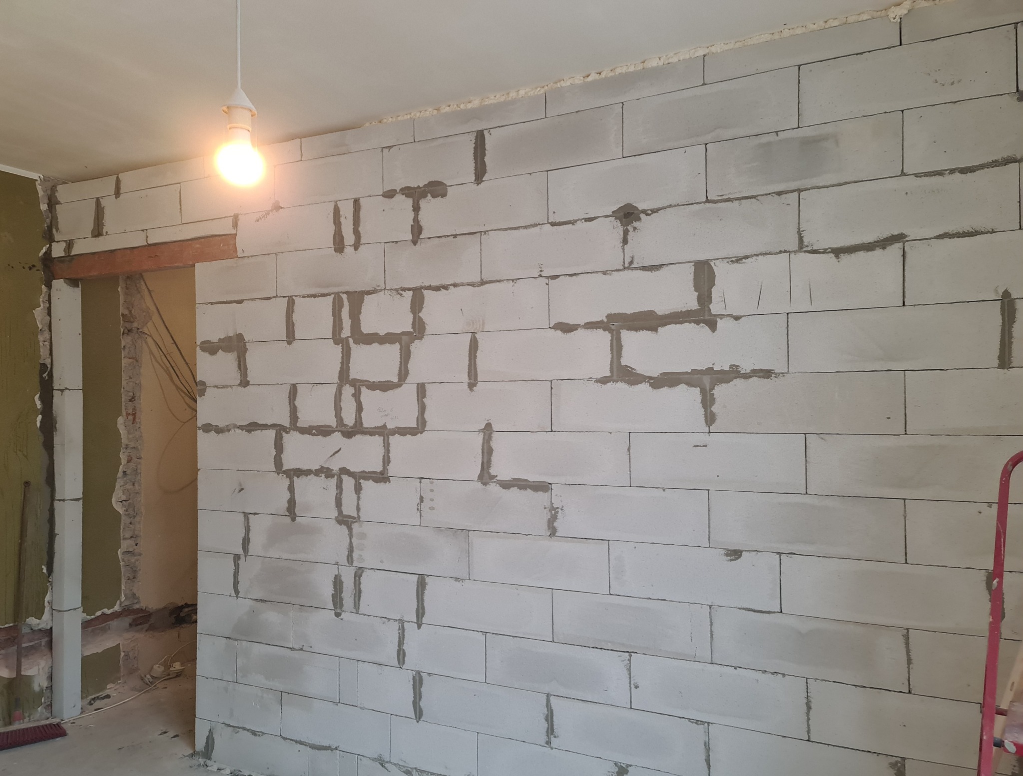 Wznoszona ściana z betonu komórkowego, widoczne spoiny i fragment otworu drzwiowego z drewnianą belką nadprożową, surowy stan wykończenia wnętrza.