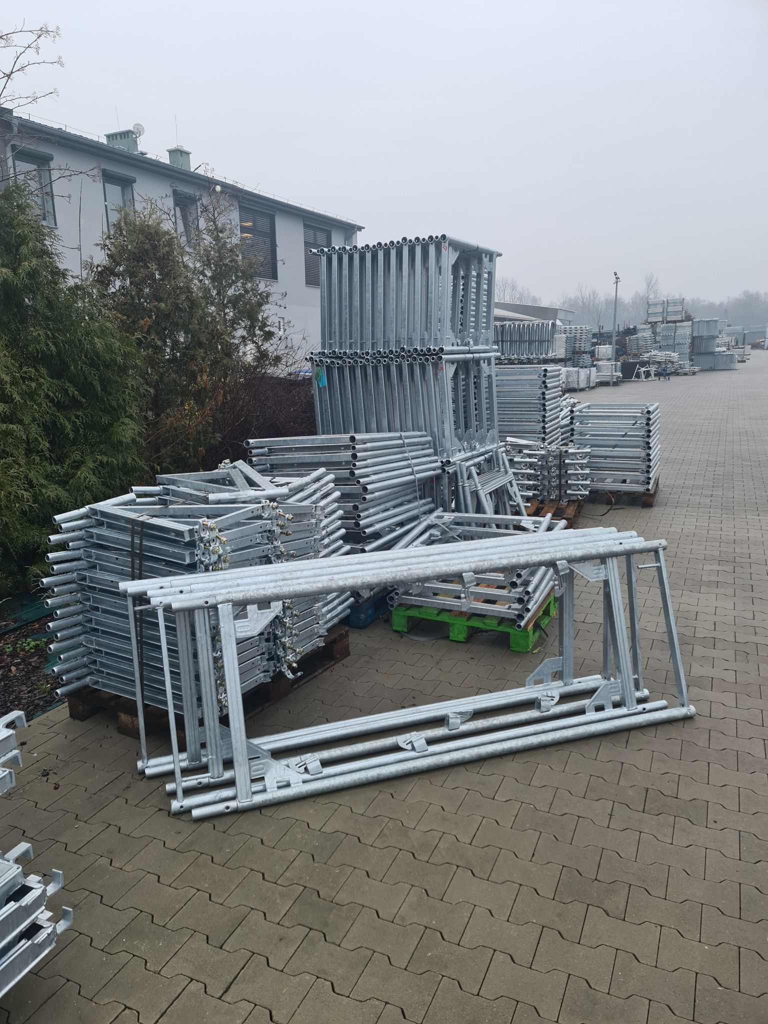 Stosy metalowych elementów rusztowań budowlanych ułożone na paletach na zewnątrz budynku w pochmurny dzień.