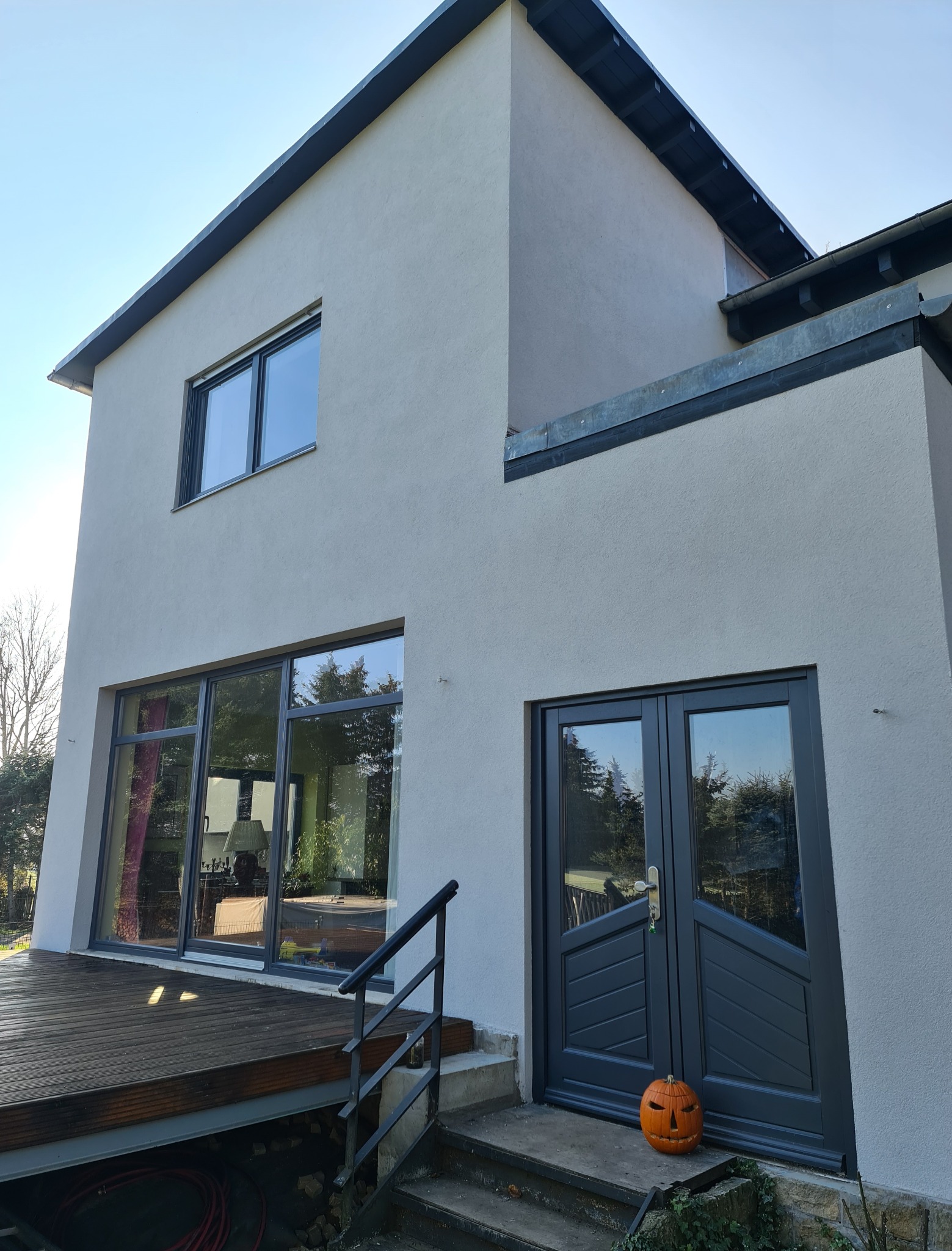Nowoczesny dom z szarą elewacją, dużymi oknami przesuwnymi i grafitowymi drzwiami wejściowymi. Na schodach przed drzwiami stoi dynia halloweenowa.