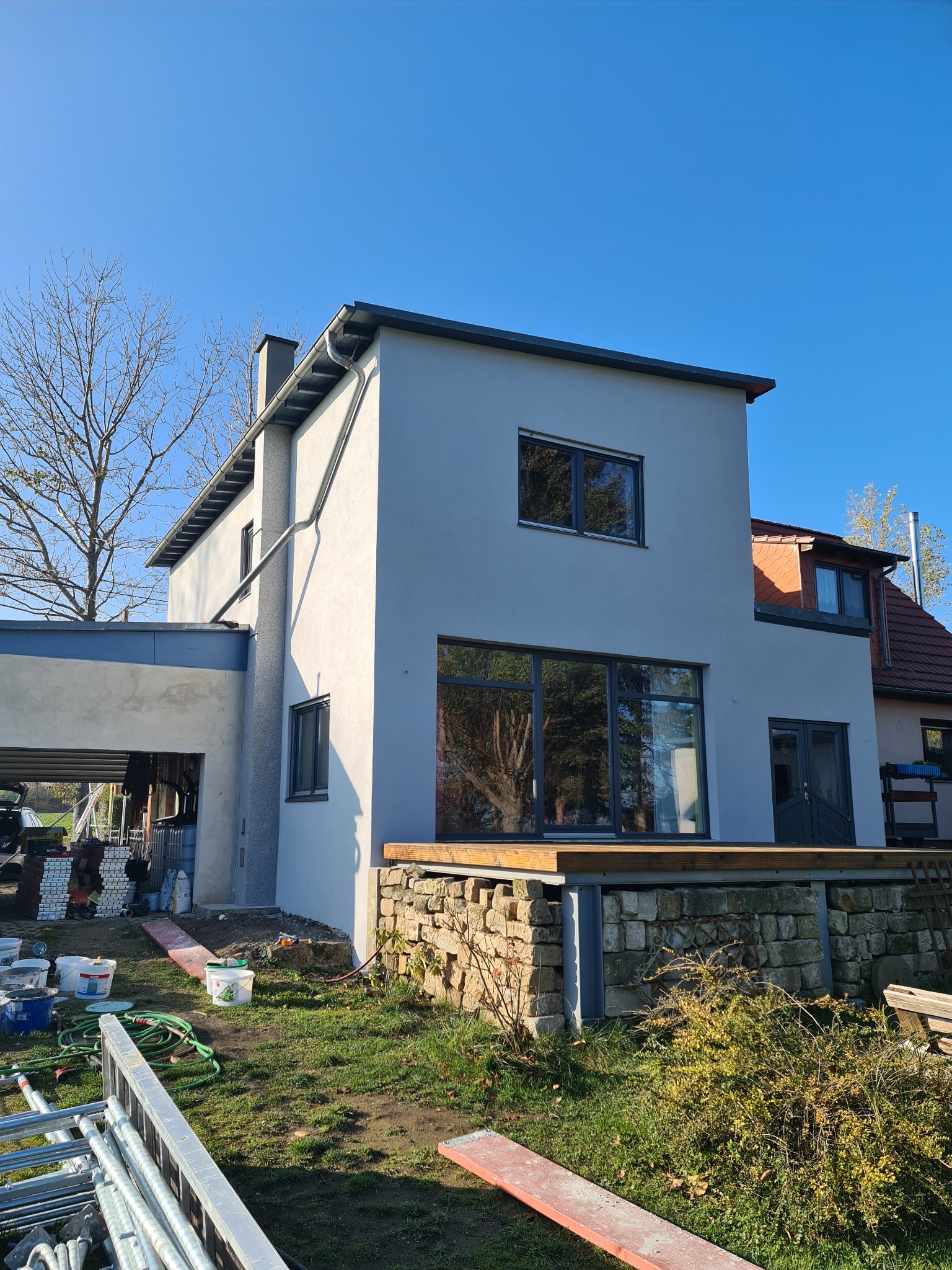 Nowoczesny dom z szarą elewacją w trakcie wykończenia, widoczne rynny, okna z ciemnymi ramami, drewniany taras i elementy rusztowania na trawniku.