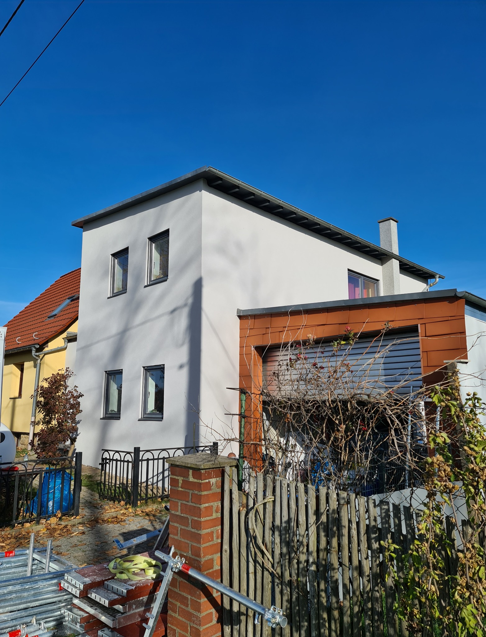 Dwupiętrowy dom z białymi ścianami, szarymi oknami i brązowym garażem, widziany zza drewnianego płotu i sterty rusztowań w słoneczny dzień.