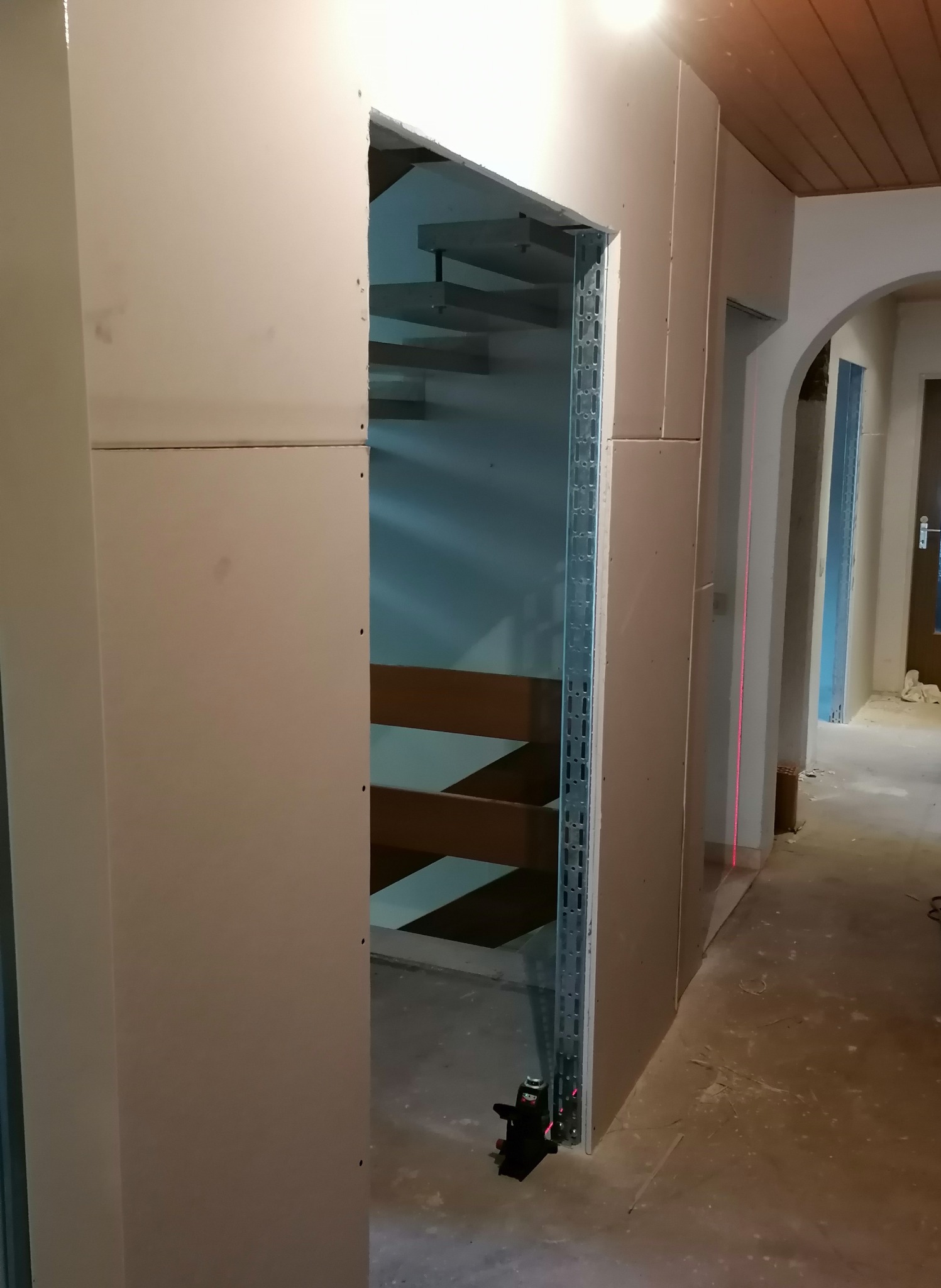 Montaż ściany działowej z płyt gipsowo-kartonowych z widocznym metalowym profilem, poziomica laserowa i fragment drewnianych schodów w tle.